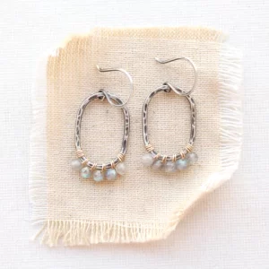 hammered silver oval hoops adorned with labradorite beads afixed with gold wire styled on tan linen.Sarah DeAngleo Jewelry box with signature gift wrap and feather styled on a yellow plate with dried grass and a crystal rock Labradorite Wrapped Mixed Metal Oval Earrings